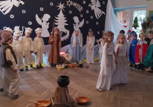 Dzieci w strojach jasełkowych stoją w półkolu, a na środku dwie dziewczynki. Przedszkolaki śpiewają piosenkę "Najpiękniejsze dni w roku".