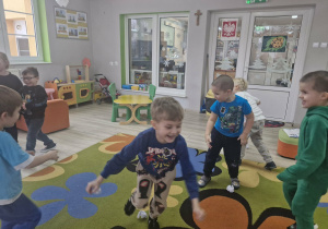 Przedszkolaki podczas zabawy ruchowej biegają po dywanie i zbierają "śnieżki" wykonane z białego papieru.