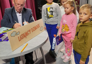 Czworo dzieci stoi obok stolika, przy którym siedzi pisarz. Autor podpisuje dla przedszkolaków książkę. Na stoliku znajduje się tabliczka z napisem "Latający Dziadek".