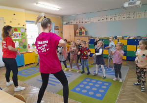 Dzieci ustawione naprzeciwko instruktorek tańca poruszają się według ich wskazówek. W tle szafa, szafki z układankami, domek dla lalek, alfabet wiszący na ścianie.