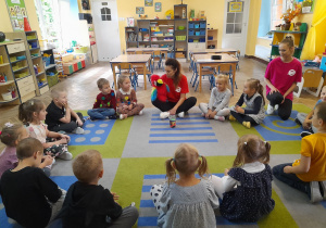 Dzieci siedzą w kole na dywanie wraz z instruktorkami Studia Tańca Alibi. Pani pokazuje dzieciom maskotkę papugi.