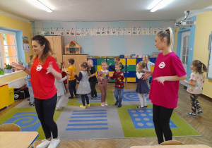 Panie instruktorki ubrane w kolorowe podkoszulki z logo Alibi obracają się wokół własnej osi, a dzieci naśladują kroki taneczne.
