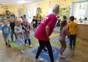 Dzieci poruszają się po obwodzie koła razem z instruktorką. W tle kąciki zabaw, tablice wiszące na ścianie, stoły, okna, drzwi.
