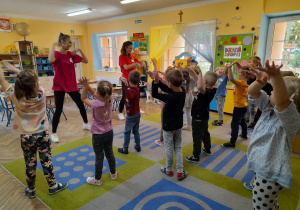 Dzieci ustawione naprzeciwko dwóch Pań instruktorek tańczą z rękami uniesionymi do góry. W tle kącik z układankami, tablice, stoliki, okna.