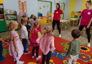 Dzieci stoją naprzeciwko instruktorek tańca i z uwagą słuchają poleceń. Panie ubrane w kolorowe podkoszulki z logo Alibi.