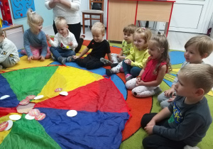 Dzieci siedzą na dywanie wokół rozłożonej chusty, na której znajdują się kropki i próbują wymyślić "Co mówi kropka?”