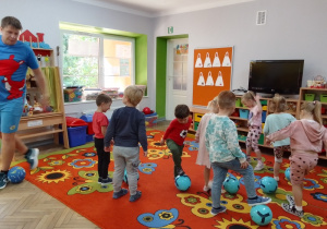 Dzieci poruszają się po dywanie z niebieskimi piłkami przy nodze. Obok - w niebieskim stroju instruktor z Drużyny Kangura.