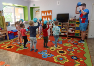 Dzieci stoją przed instruktorem, w rękach trzymają niebieskie piłki uniesione nad głowami. Przed dziećmi w niebieskim stroju stoi instruktor z Drużyny Kangura.