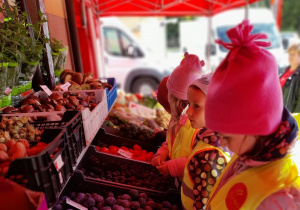 Czworo dzieci ogląda owoce i warzywa umieszczone przed sklepem w skrzynkach.