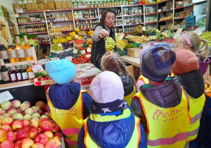 Siedmioro dzieci stoi w sklepie przed ladą, a Pani sprzedawczyni pokazuje dzieciom kiść zielonych winogron. W tle skrzynki z owocami i warzywami oraz półki, na których stoją inne artykuły.