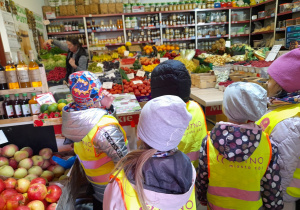 Grupka dzieci ogląda umieszczone w skrzyniach owoce i warzywa oraz inne artykuły znajdujace się na półkach w sklepie. Pani sprzedawczyni pokazuje przedszkolakom rzodkiewki.