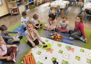 Dzieci siedzą na dywanie. Alicja zbiera z ceratki leżącej na środku dywanu śliwki i wkłada je do koszyczka. Obok stoją dwa pojemniki z marchewkami i jabłkami.