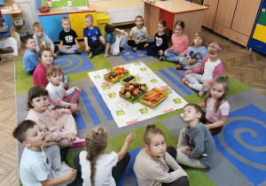 Dzieci siedzą na dywanie wokół rozłożonej ceraty, na której znajdują się cztery pojemniki z posegregowanymi owocami i warzywami.