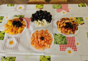 Na stole przykrytym ceratą stoją cztery talerze z rozłożonymi owocami: mandarynkami, brzoskwiniami, gruszkami, śliwkami.