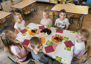 Sześcioro dzieci siedzi przy stoliku, na którym stoją talerze z rozłożonymi owocami.