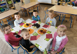 Uśmiechnięte przedszkolaki siedzą przy stoliku nakrytym ceratą. Przed dziećmi leżą serwetki, a na talerzach witaminowe smakołyki: mandarynki, śliwki, brzoskwinie, gruszki.