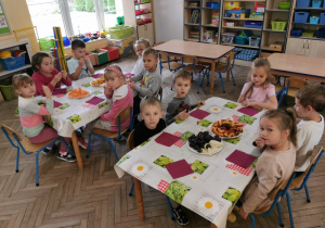 Dzieci siedzą przy dwóch stolikach nakrytych ceratą i zajadają owoce. W tle okno, dwa stoły, kąciki zabaw.