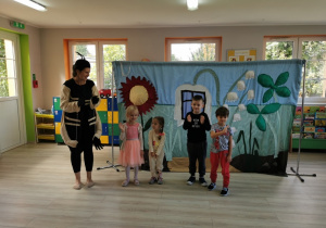 Czworo dzieci stoi na środku sceny obok aktorki przebranej za mrówkę i przedstawia się. W tle dekoracja do przedstawienia.