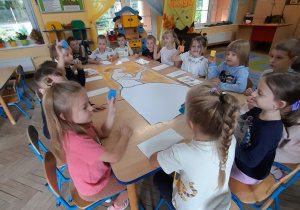"Słoneczka" siedzą przy wspólnym stole, na którym leży narysowana postać Pani Jesieni. Dzieci trzymają w dłoniach małe gąbki.