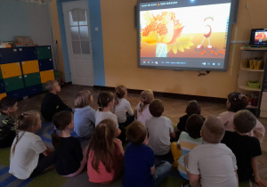 Dzieci siedzą na dywanie przed tablicą multimedialną i oglądają film edukacyjny o jesieni.