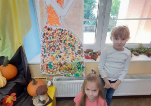 Na oknie wisi ozdobiona przez dzieci sylweta Pani Jesieni. Obok stoi Oliwia, a przed nią siedzi Vanessa. Z lewej strony fragment dekoracji z okazji powitania nowej pory roku, a na parapecie leżą szyszki, jarzębina.