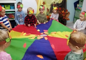Dzieci ustawione w kole trzymają chustę animacyjną, na której znajdują się kolorowe listki. W tle dekoracja z okazji Powitania Jesieni.