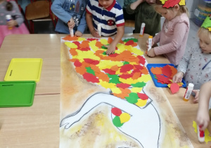 Dzieci wykonują pracę plastyczną przyklejając wycięte z papieru listki na sylwecie Pani Jesieni.