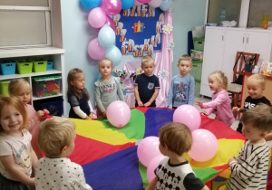 Zabawa z chustą animacyjną i balonami. Dzieci stoją w kole trzymając chustę animacyjną z balonami. W tle dekoracja z okazji Dnia Przedszkolaka.