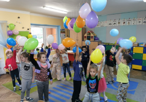 Przedszkolaki stoją na dywanie trzymając kolorowe balony w górze.