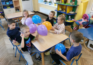 Oliwier siedząc przy stoliku pokazuje co namalował na swoim fioletowym balonie. Pozostali przy stoliku: Igor, Vanessa, Hubert, Alicja i Mieszko nadal ozdabiają swoje balony.