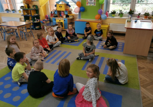Przedszkolaki siedzą w kole na dywanie podczas zabawy w rozpoznawanie rówieśników po głosie. Na środku z opaską na oczach siedzi i zgaduje Alicja.