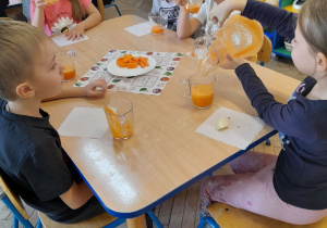 Dzieci siedzą przy stole i piją przygotowany samodzielnie sok, a Amelka wlewa z dzbanka sok do szklanki. Na środku stołu stoi talerz z marchewkami.