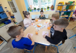 Pięcioro dzieci siedzi przy stole, degustuje sok i zajada jabłka. W tle okno, kwiaty na parapecie, kącik przyrody.