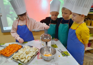 Dzieci ubrane w stroje kucharskie stoją za stołem. Amelka wyciska sok z jabłka, a Natalka, Hubert i Krzyś obserwują jak z sokowirówki wylatuje sok.