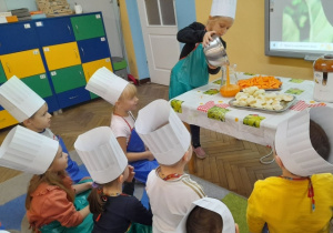 Dzieci w fartuszkach i czapkach kucharskich siedzą na dywanie przed stołem. Natalia stoi obok stołu i przelewa sok z miseczki do dzbanka. Obok leżą na tacach marchewki i jabłka.