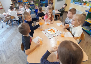 Dzieci siedzą przy stolikach, piją sok i zajadają jabłka i marchewki. Hubert dolewa sobie soku do szklanki. W tle półki z układankami.