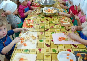 Motylki siedzą przy długim stole, na którym są miski z owocami i deski do krojenia. Dzieci ubrane w fartuszki i białe chusteczki na głowach, w rękach trzymają plastikowe noże.