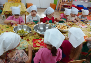 Grupa dziewczynek w kucharskich strojach kroi plastikowymi nożami owoce. Przed nimi na stole stoją metalowe miski z owocami.
