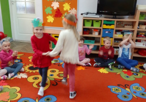 Zuzia i Julia tańczą w parze w środku koła z opaskami z liści na głowach, reszta dzieci siedzi na obwodzie koła i klaszcze w ręce.