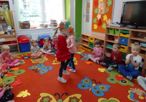 Zuzia i Oliwia poruszają się w parze w rytm muzyki z opaskami na głowach, dzieci na dywanie klaszczą w ręce. W tle kąciki zabaw, tablica z kolorowymi liśćmi.