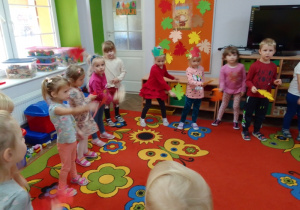 Dzieci poruszają jesiennymi liśćmi na tle piosenki "Kolorowe listki". Z tyłu: dekoracja na tablicy, telewizor i kąciki zabaw.