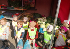 Dzieci ubrane w kamizelki odblaskowe pozują do zdjęcia przed sklepem owocowo-warzywnym. W tle skrzynki z owocami i warzywami.