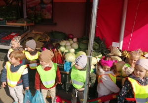 Dzieci stoją na chodniku przed sklepem owocowo-warzywnym i trzymają węża oraz zakupione owoce. W tle skrzynki z owocami i warzywami.
