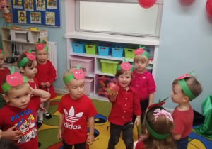 Dzieci stoją w rozsypce na dywanie, ubrane są na kolor czerwony, a na głowach mają opaski z jabłkami. Na środku stoi Staś, który trzyma w ręku jabłko. W tle wiszą czerwone balony z zielonymi listeczkami.
