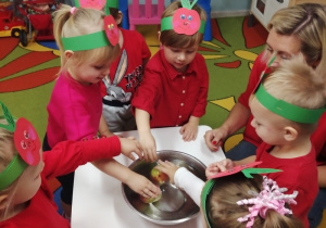 Dzieci stoją wokół białego stolika i badają "czy jabłka potrafią pływać". Na stoliku stoi miska z wodą, do której przedszkolaki wkładają jabłka.