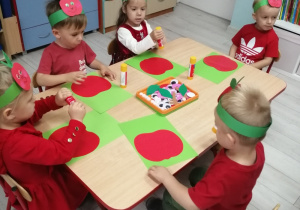 Grupka dzieci siedzi przy stole podczas wykonywania pracy plastycznej. "Pszczółki" naklejają na zielony karton czerwone jabłko.