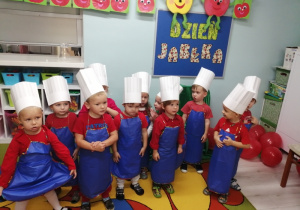 Zdjęcie grupowe dzieci, które ubrane są w granatowe fartuszki oraz białe czapki kucharskie. W tle dekoracja z okazji Dnia Jabłka.