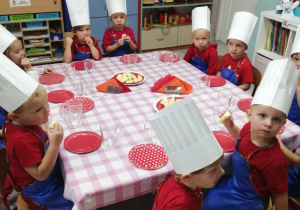 Dzieci siedzą przy wspólnym stole przykrytym ceratką, na którym znajdują się czerwone talerzyki z kawałkami jabłek, szklanki oraz serwetniki. Przedszkolaki ubrane są w granatowe fartuszki oraz białe czapki kucharskie i degustują jabłka oraz własnoręcznie wyciśnięty sok.