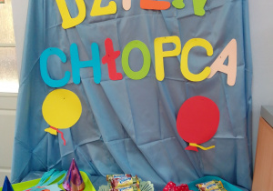 Dekoracja w sali: na tablicy napis – Dzień Chłopaka i papierowe balony, pod tablicą leżą kolorowe krawaty, słodycze i upominki.