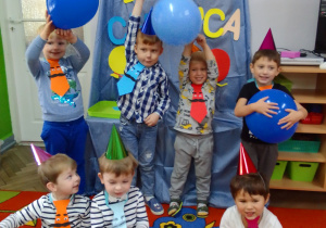 Chłopcy w kolorowych krawatach pozują do zdjęcia, w rękach trzymają niebieskie balony, w tle dekoracja.